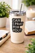 Cream-colored tumbler with 'Can I Pray For You?' text on a wooden surface with plants and a candle in the background.